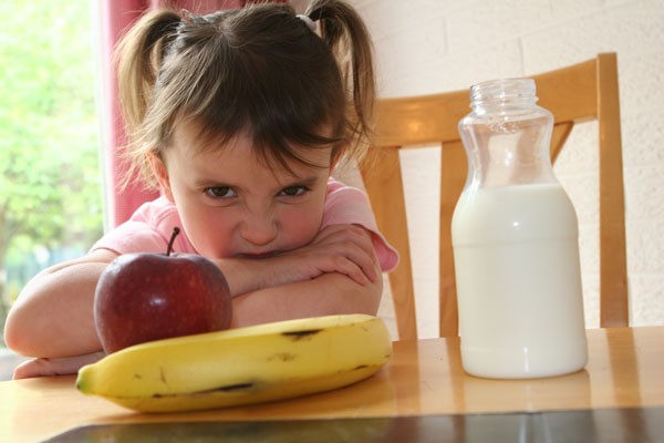 An upset child sitting at a table with fruit and milk.