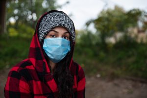 Sad young girl or woman with hood and protective medicine mask standing outside.