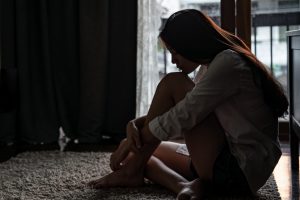 Young woman sitting on bedroom floor hugging her knees and thinking.